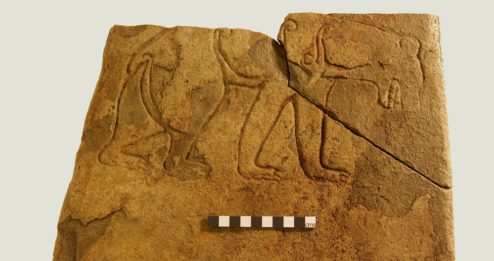 A yellow stone slabe with a large crack across the top right corner. On the top two thirsd of the slab is a carved bear, with its head crouched downwards.