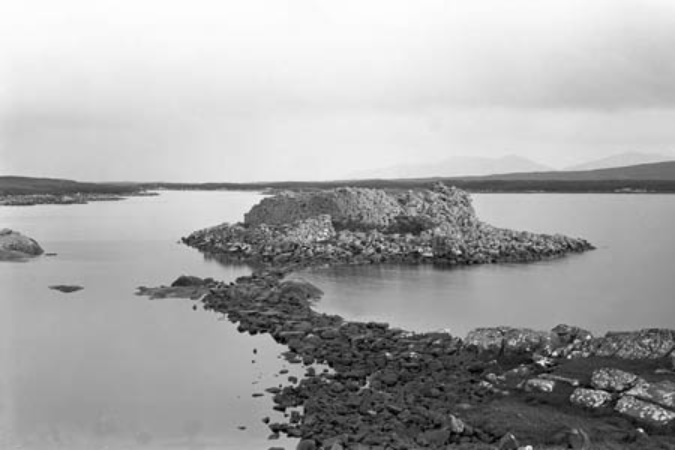 Black and white image of a rocky island in a low body of water, attached to the land in the foreground by a thing strip of dark rocks and earth. 