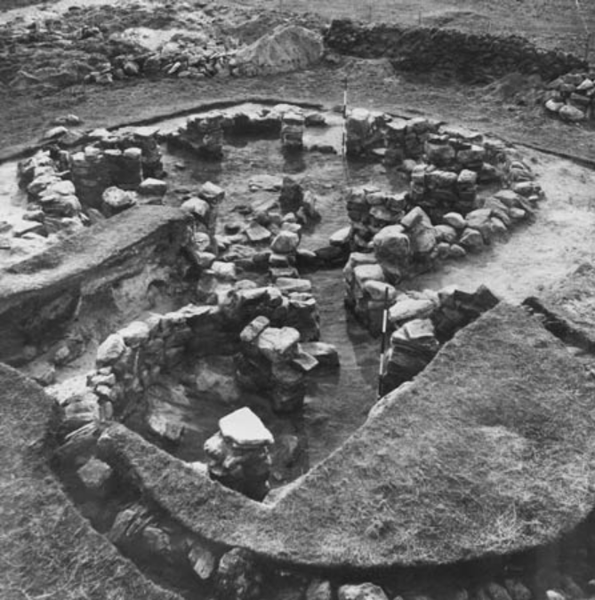 Black and white photograph of a stone structure made up of two sepaprate areas with curved walls and a connecting passage in between.