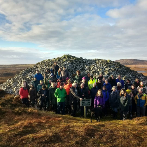 A group of smiling people pose together in front of a large mound of stones. They are all wearing 