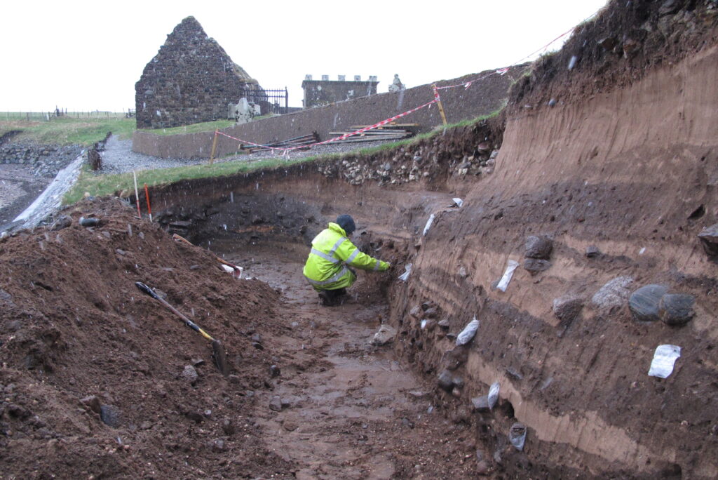 A person in a high visibility jacket crouched in a large, long trench with a clear section along one side. Sample bags are pinned into this side all the way along, and sedimentary layers are clear.