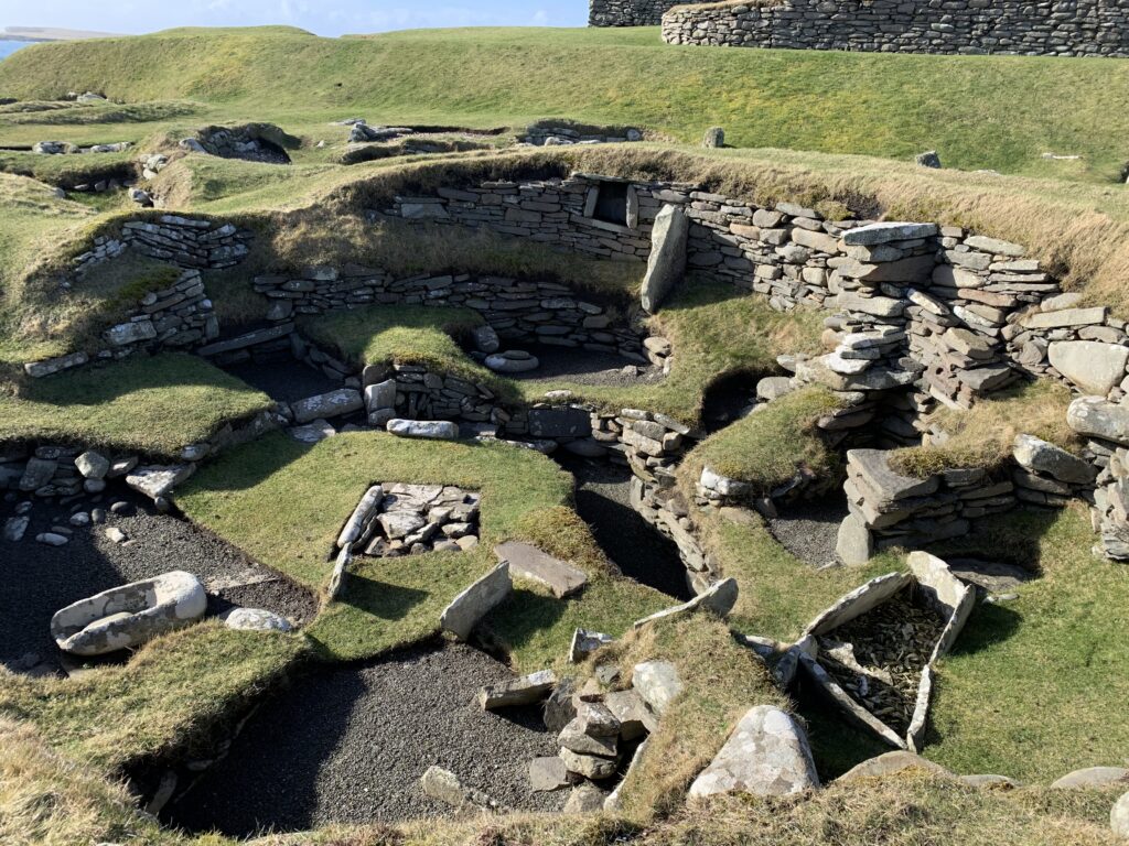 Looking obliquely down into a stone and turf building in excellent condition, showing the hearth in the centre. 