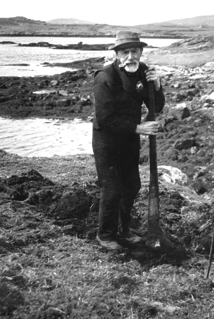 Black and white photograph of a man standing on a patch of disturbed earth on a rocky coast. He is holding a long farming tool. He is wearing a hat and has a pipe in his mouth.