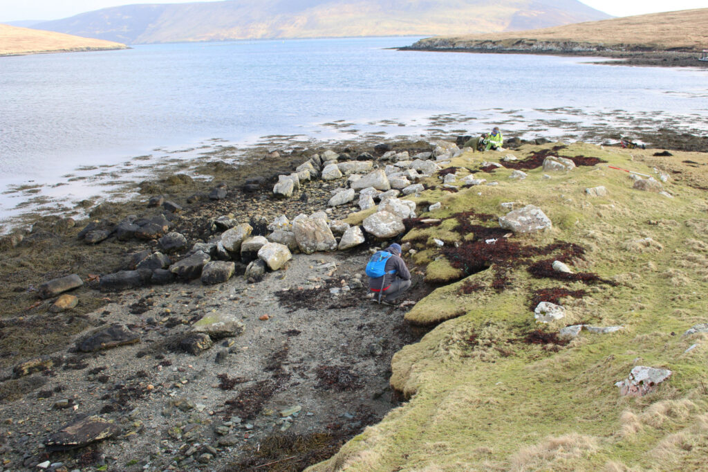 A person in blue and grey waterproofs crouched down on a rocky coastal area between a grassy area and the sea. Large boulders clustered together appear to be an ancient building as risk of being taken by the tide. 