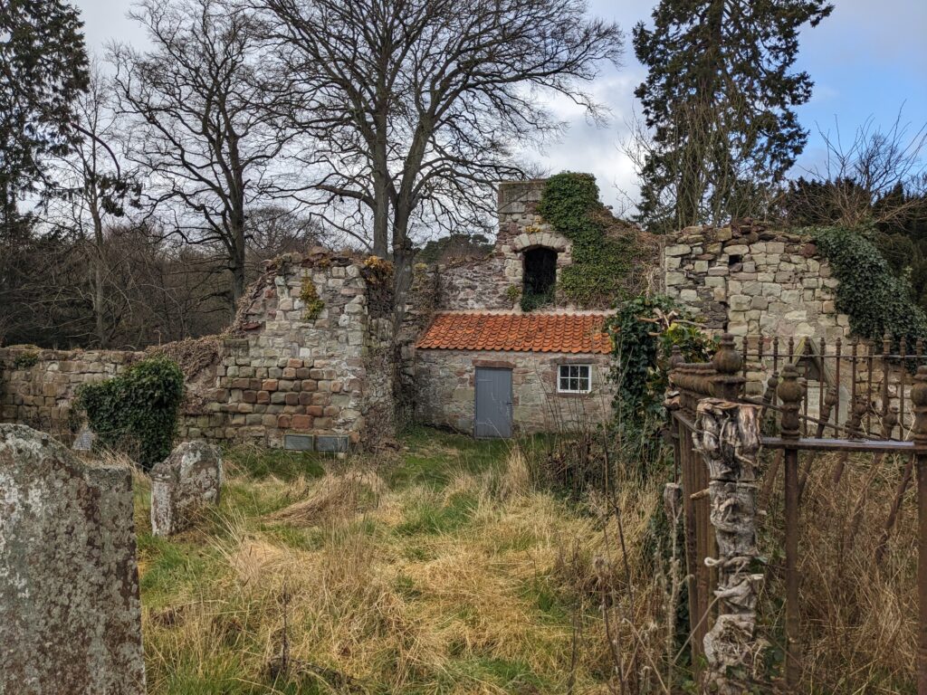 Ruined stone church walls, with small stone building at back interior wall, surrounded by long grass