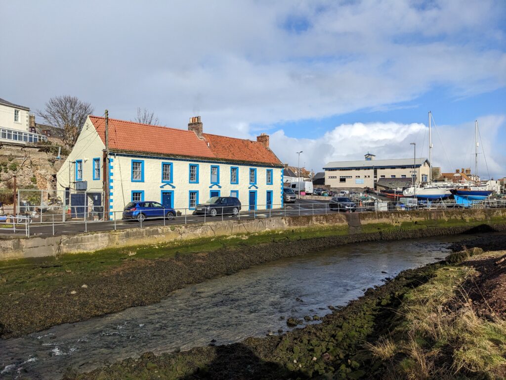 Photograph of harbour with river in front of white building and road, and boats to the right
