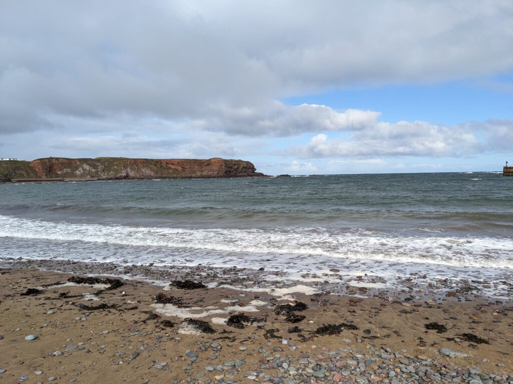 View of a headland protruding into a calm blue sea, with sand at the forefront