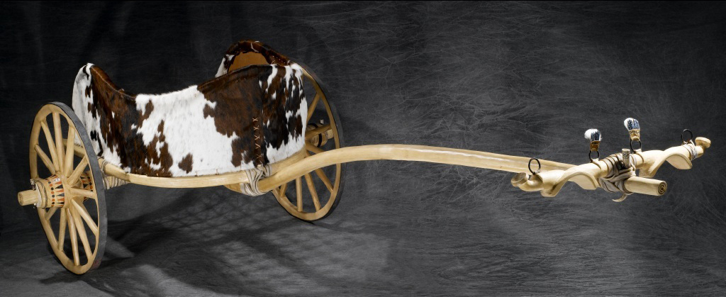 Photo of a cowhide and wood chariot against a dark grey background. 