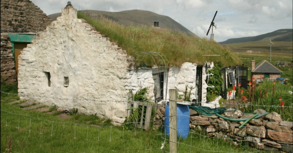 A white cottage with a turf roof and a drystone surrounding wall