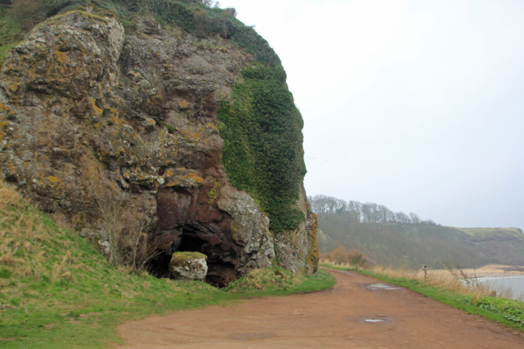 Image of the outside of a cave in the side of a steep, green and red cliff. 