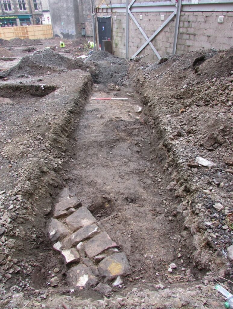 photograph of excavations, with rectangular trench. Feature of linear stone wall at closest edge of trench