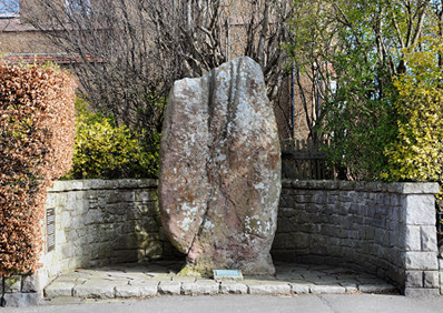 Standing stone in an alcove off of the pavement and surrounded by hedges