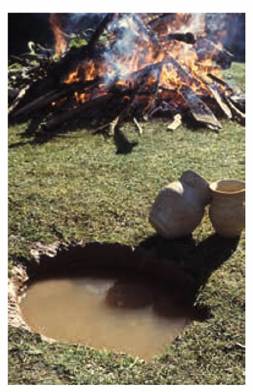 Photograph of pit with two pots on the edge in front of a small fire