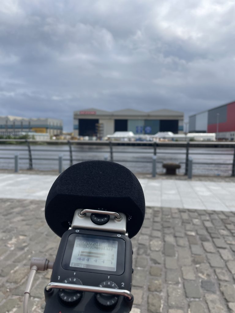 A recording device is held at the bottom of the frame, with the River Clyde seen out of focus in the background. 