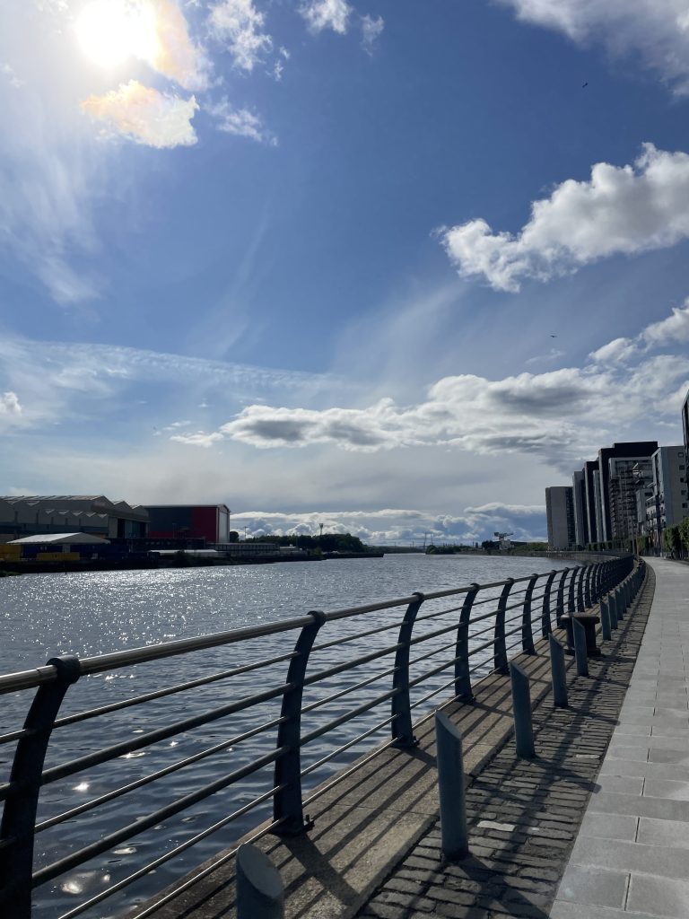 A view along the River Clyde with the sun shining in the blue sky, reflecting brightly in the water. 