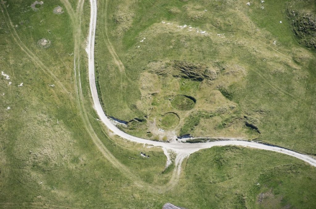 Oblique aerial view of a green, grassy landscape with two clear oval indentations in the land, looking semi-submerged nut still grass-covered.