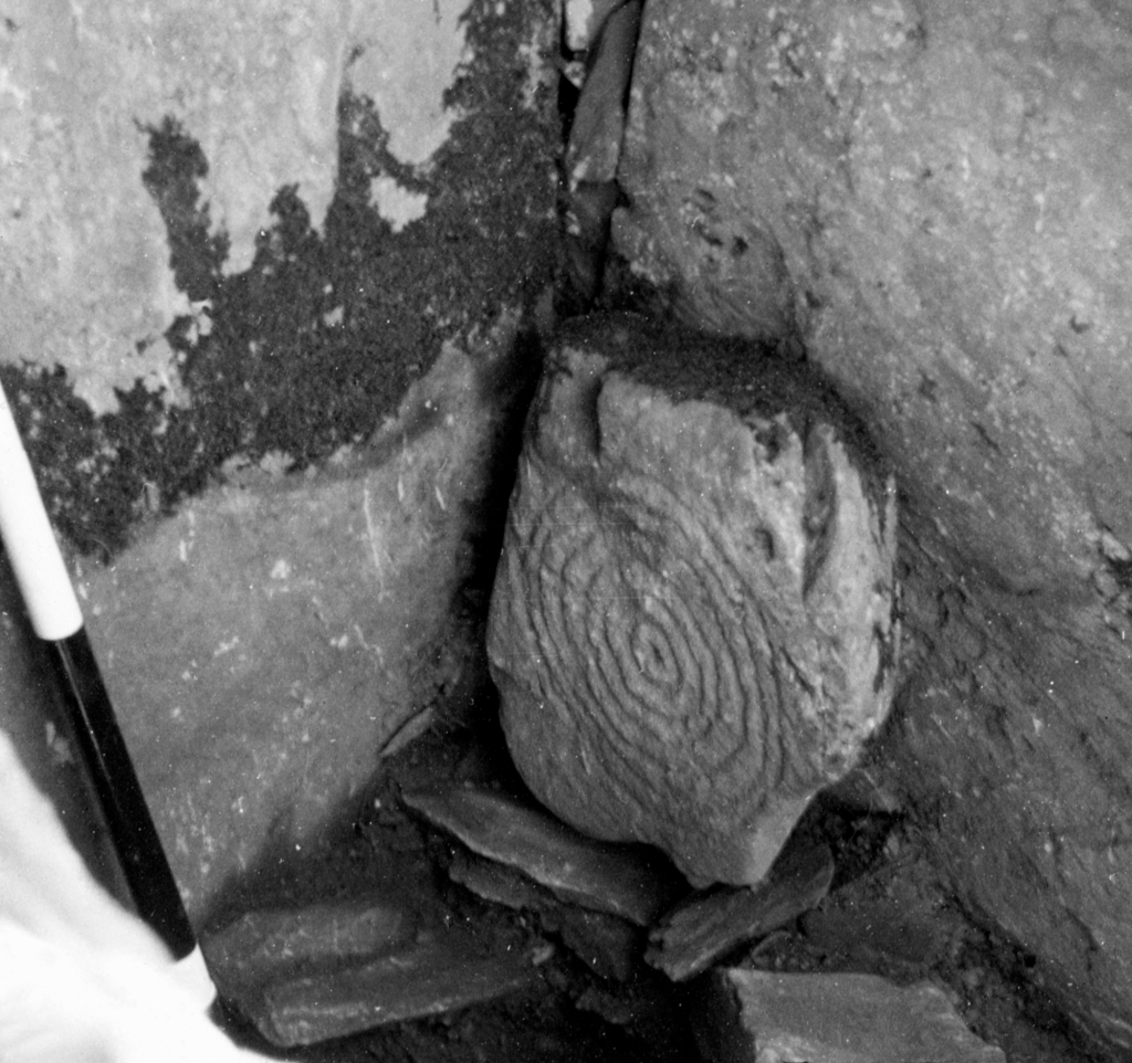 Black and white photo of an upright stone in the corner of a stone building with a large spiral motif.