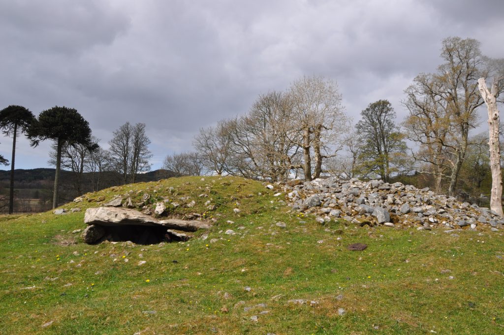An external view of a cairn which resembles a grass covered hill with some exposed stones and one large protruding slab casting a shadow across an apparent entrance. 