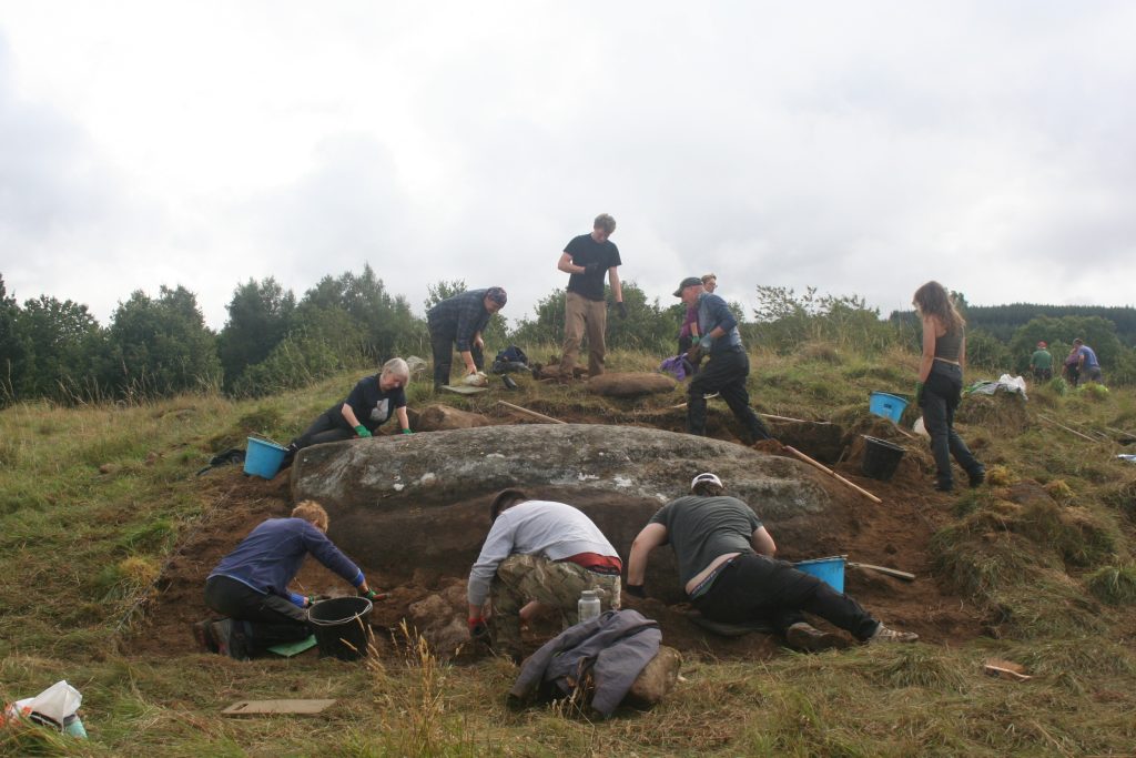 An active excavation of a large stone in a grassy hillside. They are scraping back the soil and crouched all around the large sloping stone.
