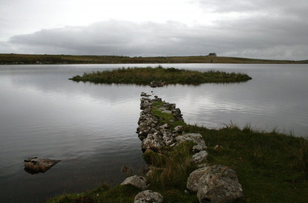View across a loch showing a stone8 pathway which is partially submerged. A grassy island sits in the middle of the loch. 