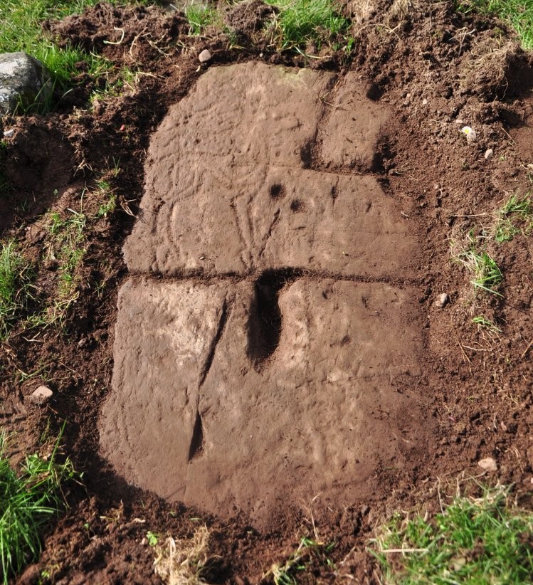 A recently uncovered, soil covered stone in the ground with a deer carved into the top left corner.
