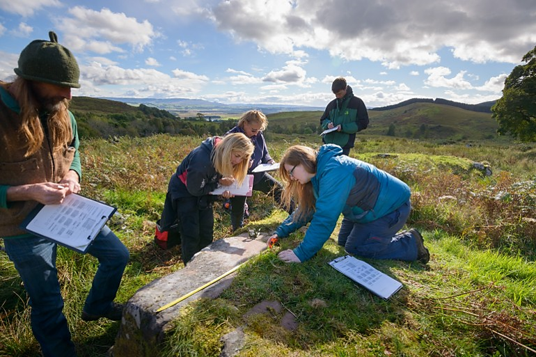 A group of five people recording a large flat stone, one measuring with a measuring tape and the others holding clipboards and pens. 