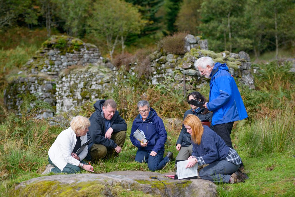 Six people kneel and bend towards a large flat stone exposed in a grassy area. One of them is touching the stone, and some of them have clipboards.