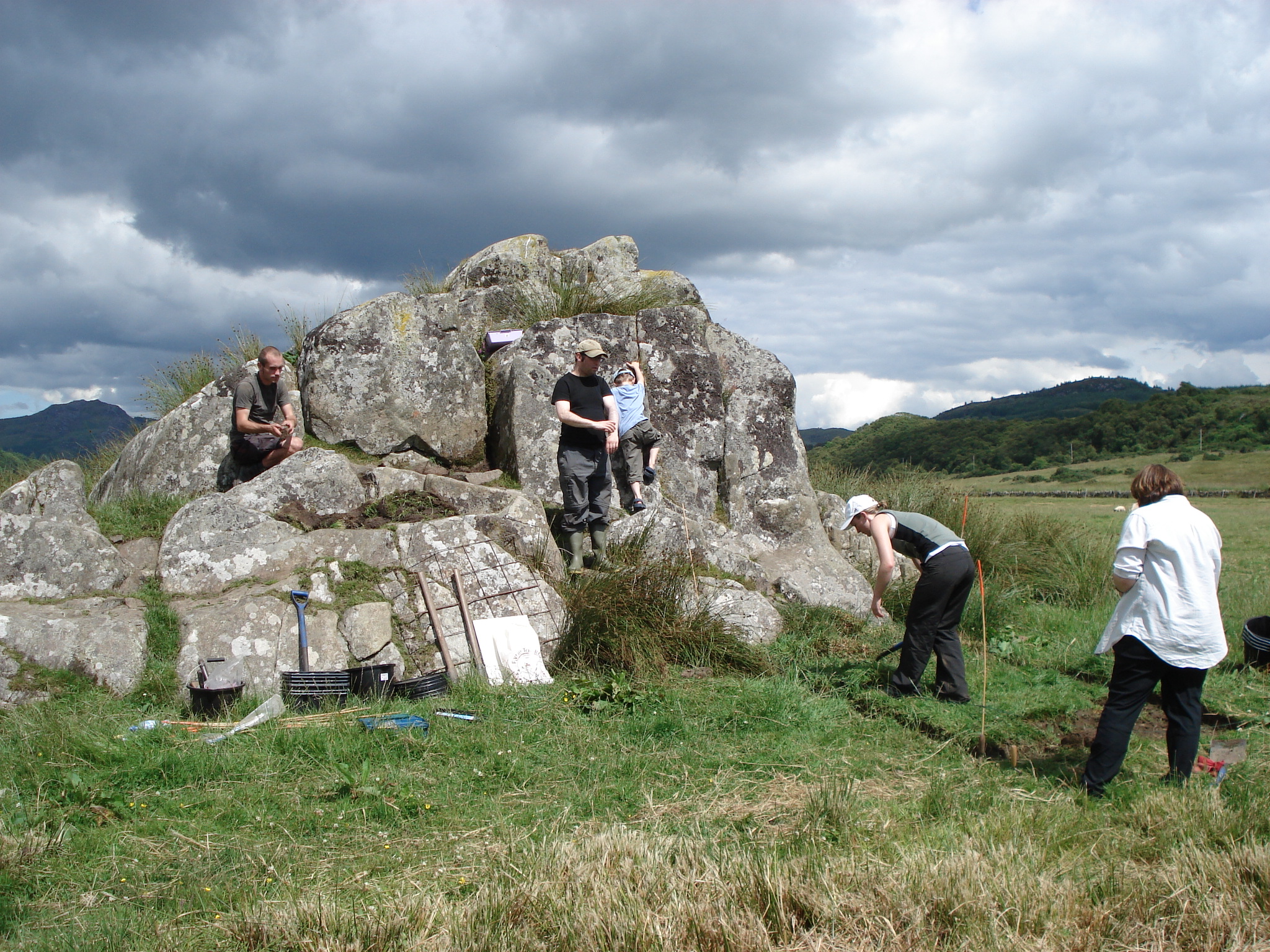 Five people working in a grassy area next to a large stone mound. Two of them are deturfing an area of grass in the foreground while the others are standing on the stones above them. 