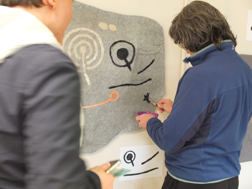 A person in a blue sweatshirt painting rock art motifs on a piece of grey felt using black paint. Other motifs can be seen in white and orange.