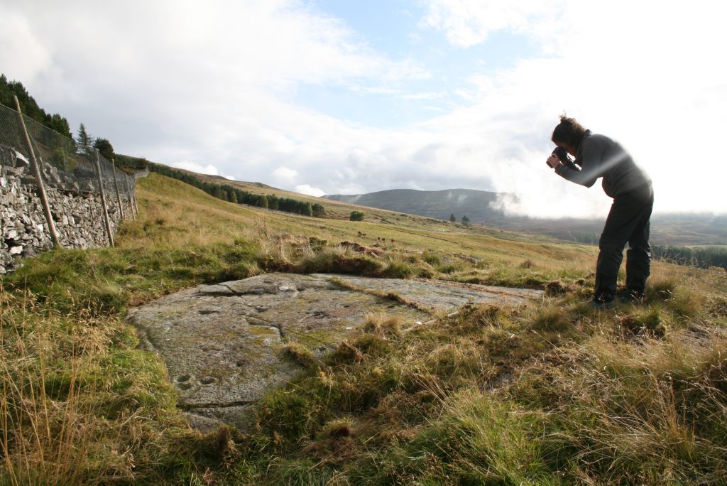 A person holding a camera to their eye and standing over a large flat rock in a grassy landscape, leaning forwards to take the picture. 