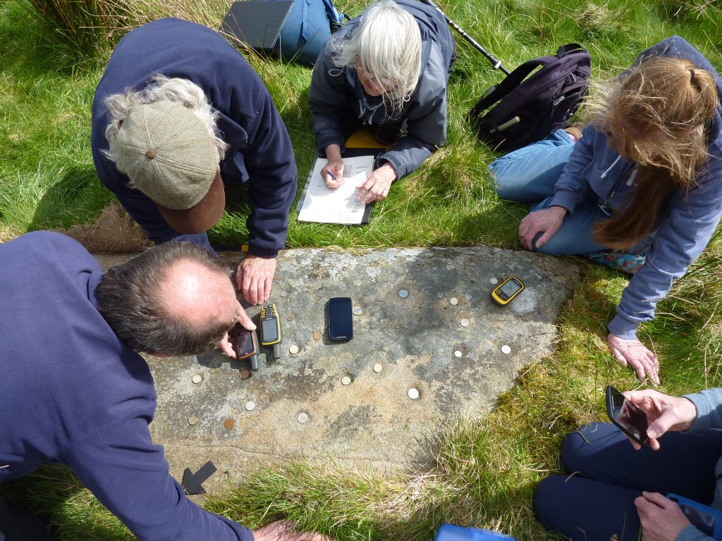 An aerial view of five people crouched over a flat stone in a grassy area, with coins placed in multiple cupmarks. One of them is lying on their stomach and sketching the stone.