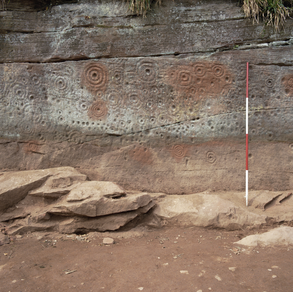 Image of a red and brown sandstone vertical cliffside with spiral motifs carved across it. 