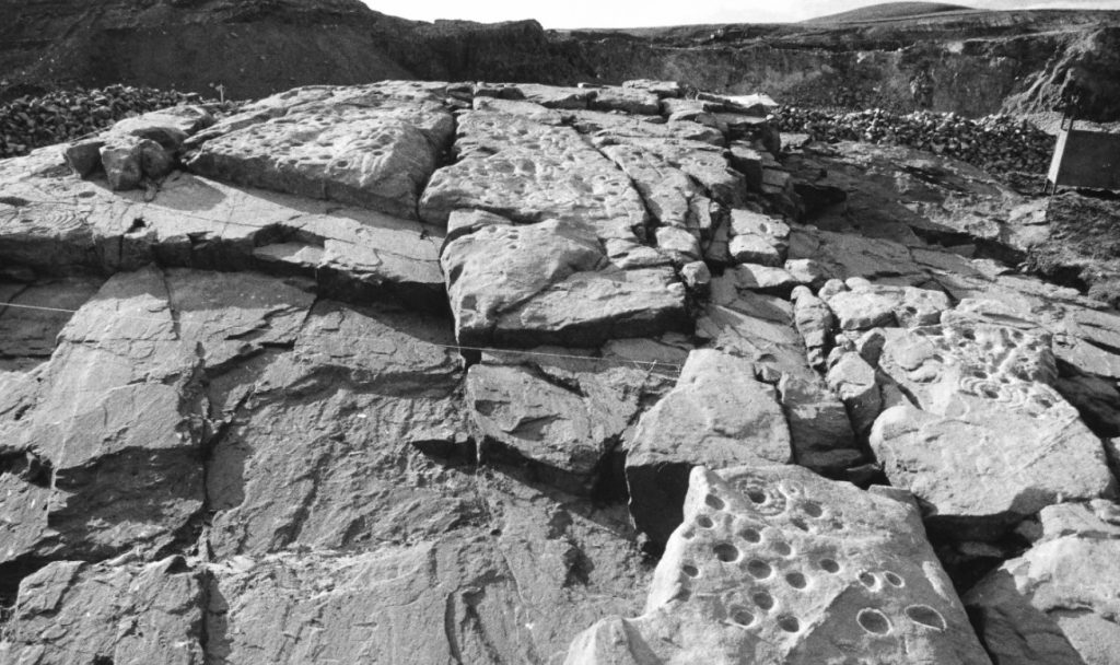 Black and white photo of a a very large and uneven outcrop of stones with various carvings on them. 