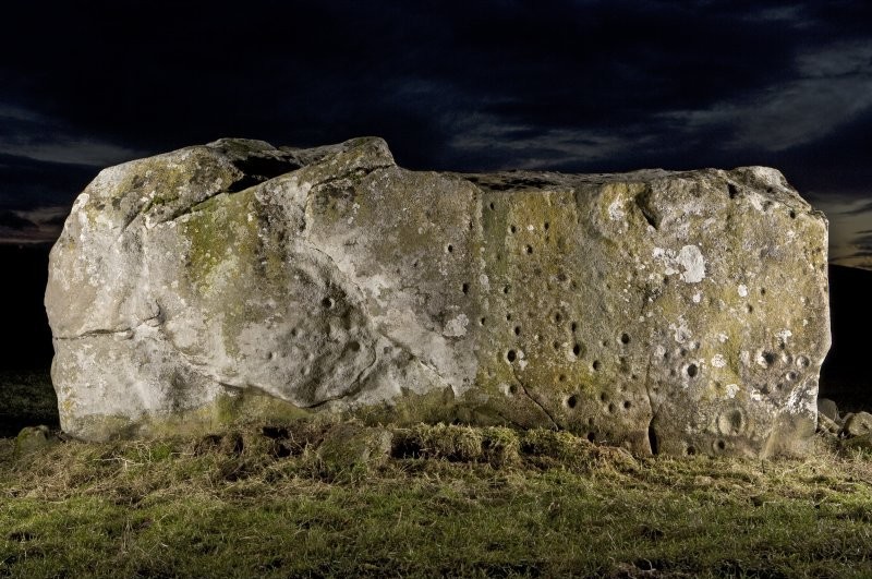 A large horizontal sub-rectangular stone on a grassy ground with cupmarks and circles on the side. Image taken against a dark night's sky. 