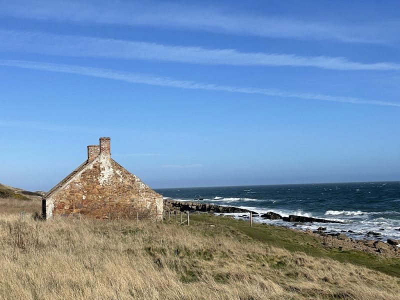 260714_NE_CrailSalmonFishermansHut On the left of the image is the gable end of a house in orange-brown stone, on the right of the image the sea is breaking over rocks. In the foreground are yellowing grasses