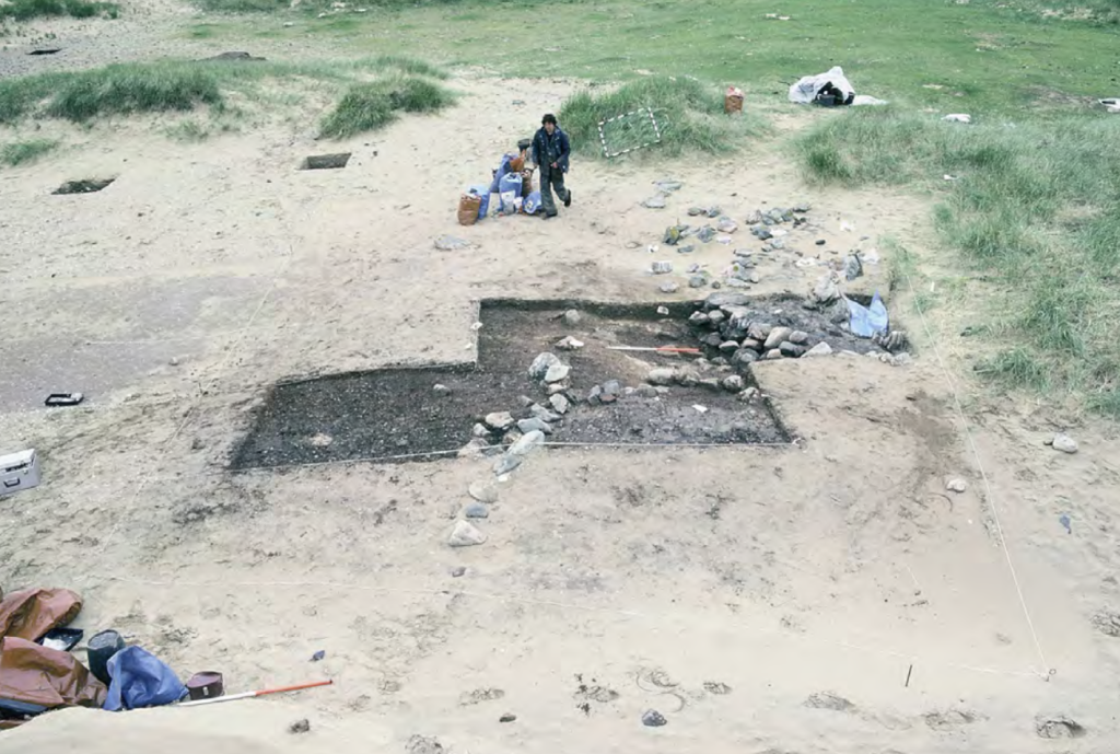 Aged photograph of an open excavation trench in a sandy area with digging equipment and a person walking in the background. 
