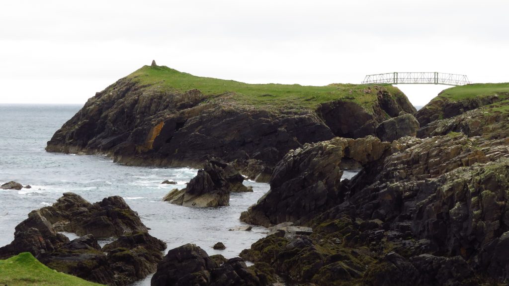 Landscape photograph showing a rocky cliffside with grass covering the top of the land. A small bridge can be seen connecting two areas of land across the rocky cliff and water. 