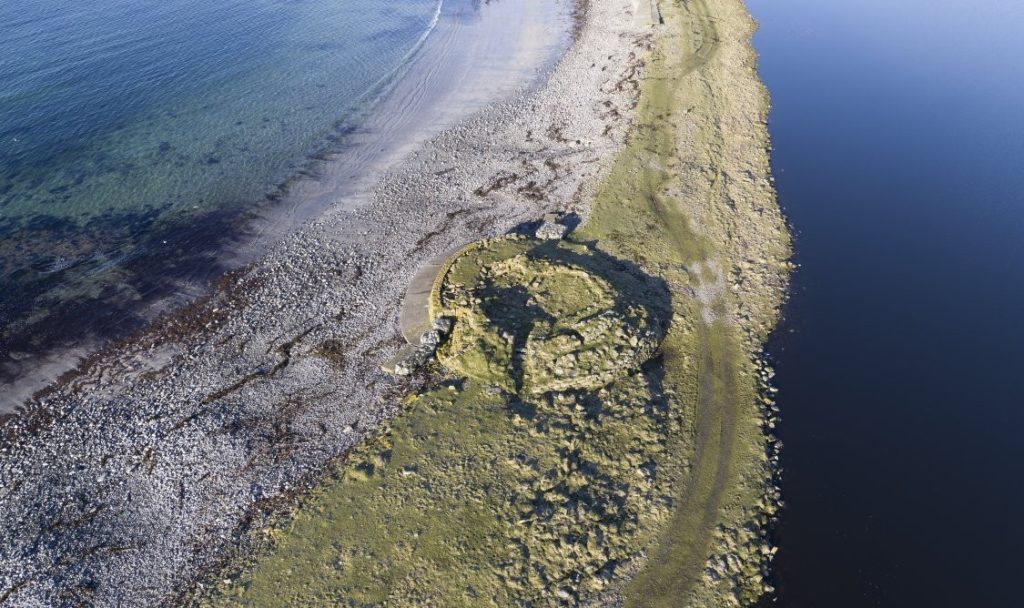 Oblique aerial view of a circular stone structure sitting on a thin strip of land with water on either side.