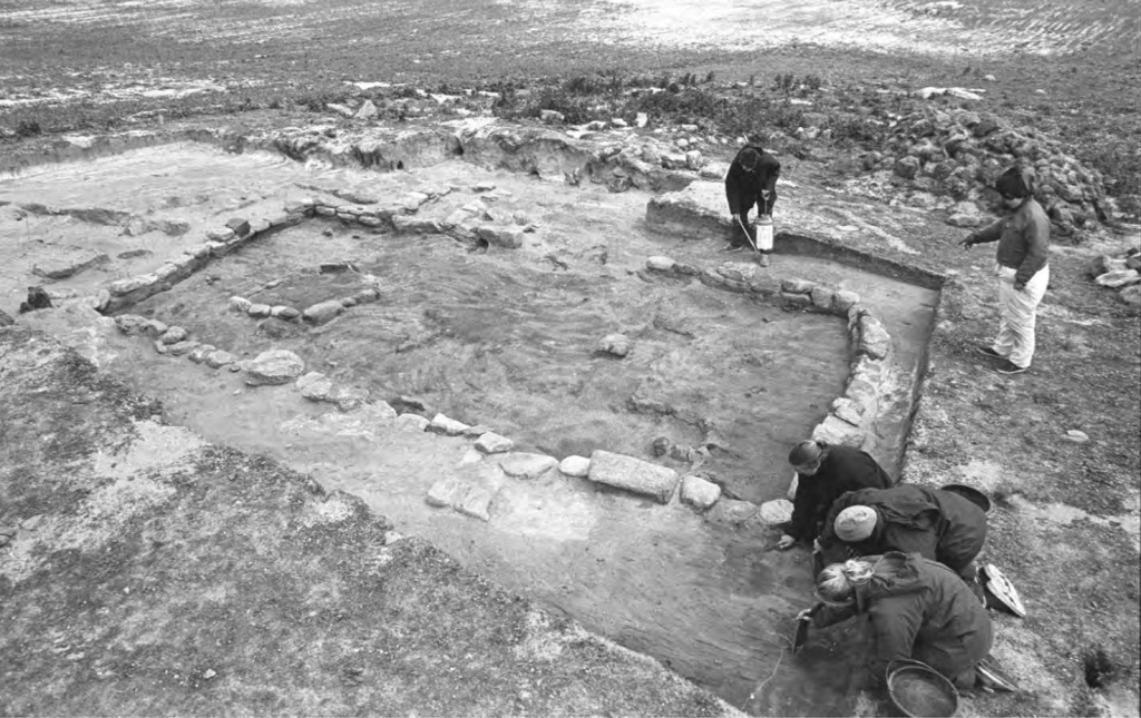 Black and white excavation photo showing a rectangular stone structure being excavated by five people. 