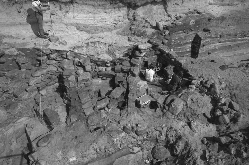 Black and white photograph looking over an active excavation site of a large stone structure. Two people are sitting on the ground inside a walled section of the structure, and another can be seen standing with a large scale pole in the background.