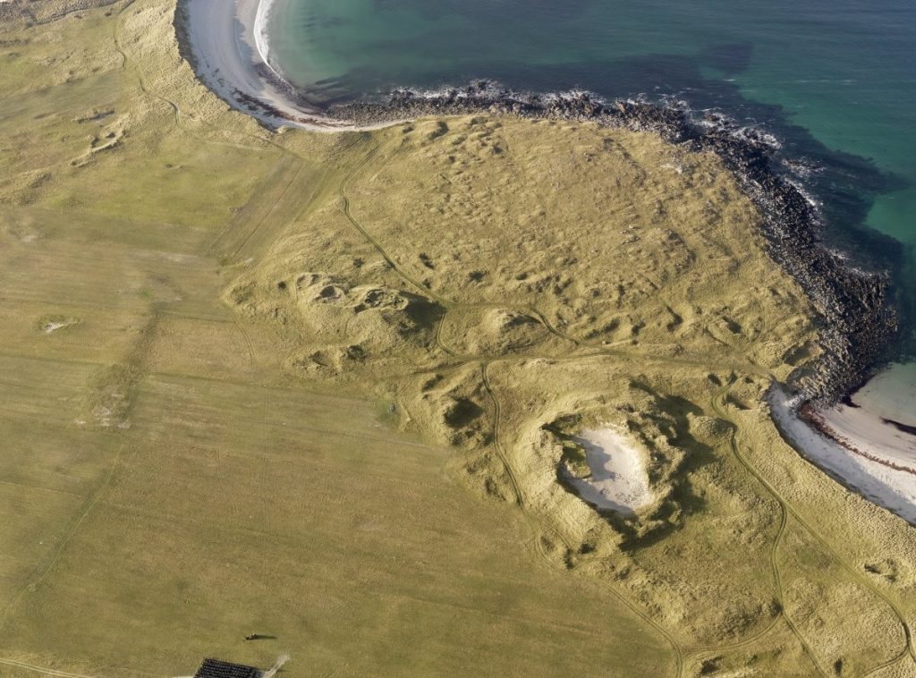 Oblique aerial photograph of a yellow-green coastline, a thin strip of white sandy beach and green-blue ocean. Archaeological features, irregular bumps in the land, can be seen across the grassy land. 