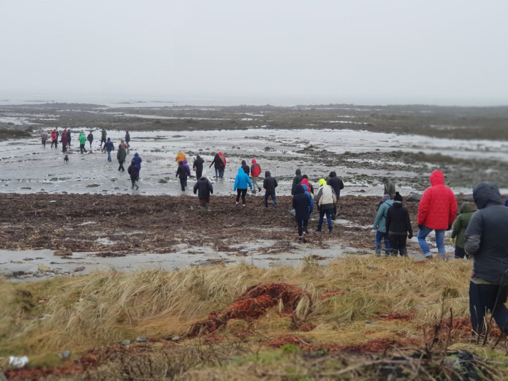 A large group of people walk in a long line across wet peatland. They are wearing waterproofs and have their hoods up because it is a rainy and windy day.