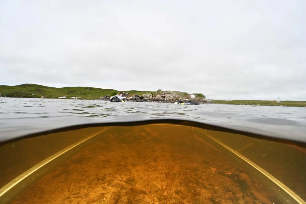 An image taken half submerged in a body of water, where the bottom half shows the metal grid under the water and the upper half shows a stone artificial island with people kneeling on it.