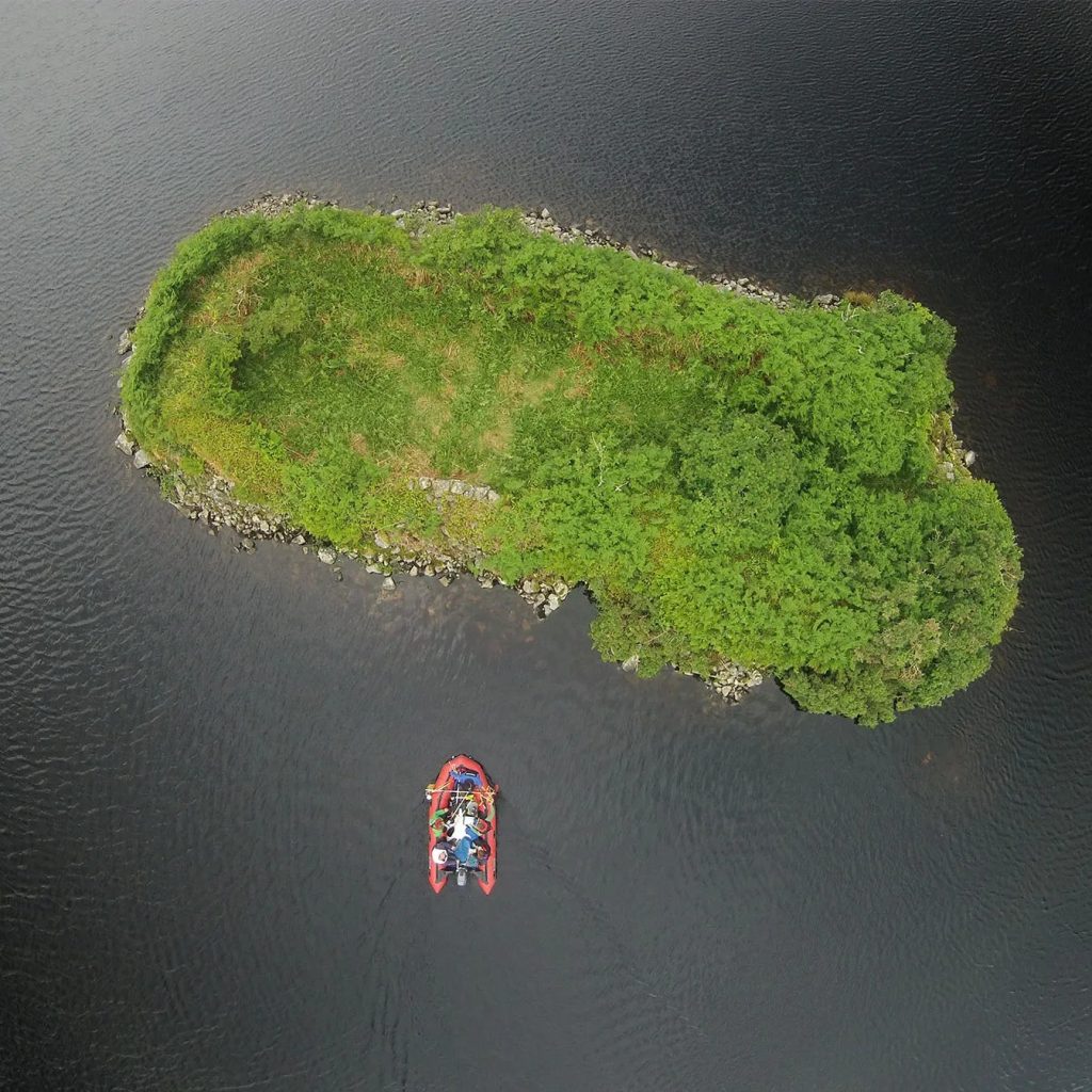 Aerial view of a green, grassy oblong island in a body of water. A small red inflatable boat can be seen to the bottom of the image, travelling towards the island.