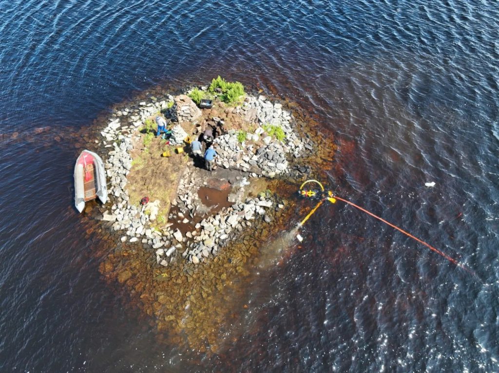 Aerial view of a stone island in the middle of a body of water. Four people can be seen on the island, digging with trowels. An inflatable boat rests at the edge of the island. 