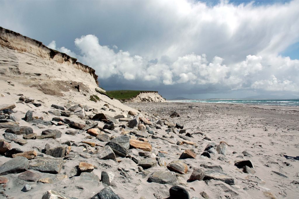 Photograph of an eroding cliffside on a sandy beach. Large stones are protruding from the cliff. 