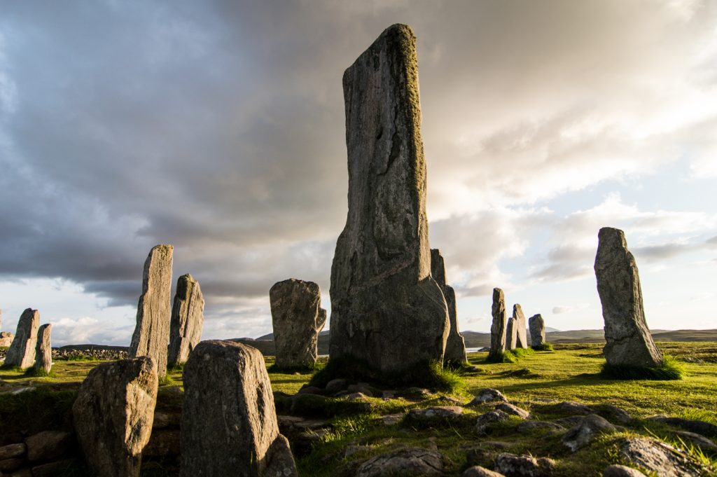 14 standing stones of different heights and shapes stand on a grassy field on a birght, cloudy day. 