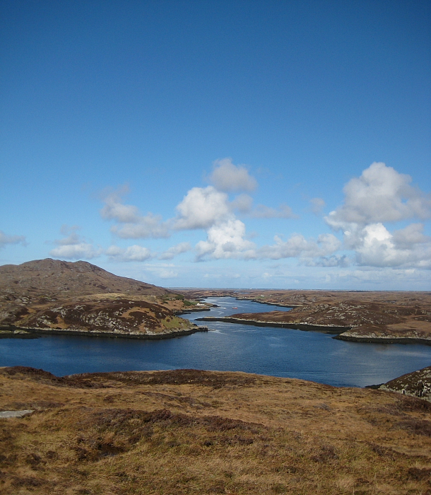 Photograph of a loch set in a vast landscape of brown, mossy land. The water is bright blue and the sky is cloudy and blue. 