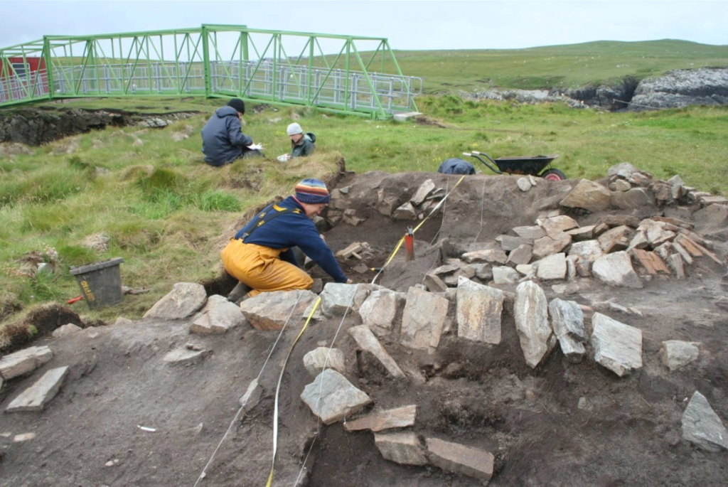 Four people excavating a large stone filled structure in a grassy area next to a small metal bridge 