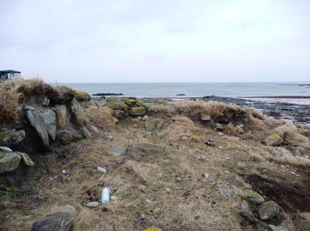 Photograph of an eroding stone structure from a pale brown peat covered patch of landc. A body of water can be seen in the background. 